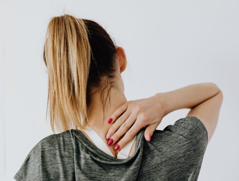 Back view of blond faceless lady in casual t shirt and manicure demonstrating pain spot on back while sitting near white wall
